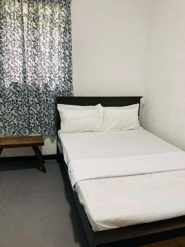 This image shows a simple bedroom with a neatly made bed, white bedding, patterned curtains, a wooden bench, and plain white walls creating a minimalist look.