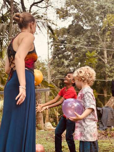 A woman and two children with balloons are playing outside. The children are smiling and interacting, surrounded by trees, creating a joyful and lively atmosphere.