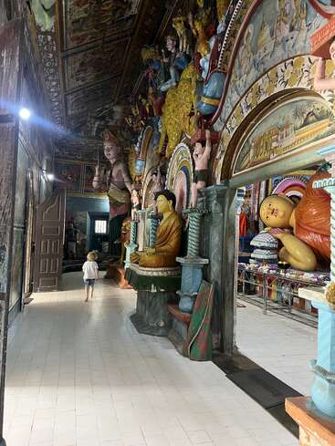 A child walks barefoot inside a vibrant temple adorned with colorful Buddha statues, detailed wall and ceiling paintings, and rich cultural decorations, creating a serene atmosphere.