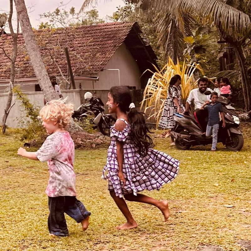 Two children joyfully run barefoot on grass, while adults and kids watch from scooters nearby. Trees, a house, and tropical plants frame this lively, heartwarming outdoor scene.