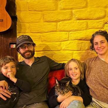 A family of four sits on a sofa in front of a yellow brick wall, smiling together. One child holds a cat. A guitar hangs above.