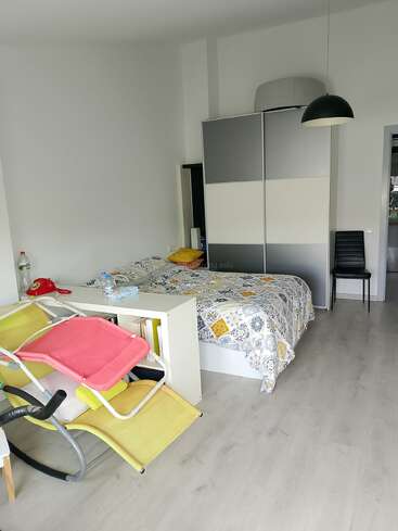 This image shows a modern, minimalistic bedroom with a double bed, colorful lounge chair, wardrobe, pendant lamp, chair, books, and various everyday objects.