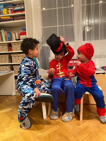 Trois jeunes enfants sont assis ensemble à l'intérieur, vêtus de costumes colorés : l'un dans un costume d'animal bleu, l'autre en maître de cérémonie et le troisième en Spiderman, et interagissent joyeusement.