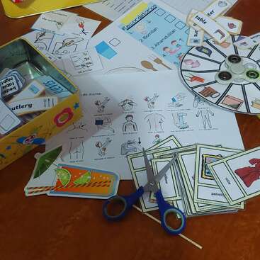 Esta imagen muestra materiales educativos, como tarjetas ilustradas, hojas de trabajo, una ruleta, etiquetas de palabras y tijeras, organizados para una divertida actividad interactiva de aprendizaje o de idiomas.