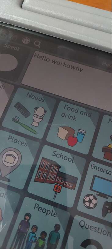Cette image montre l'écran d'un dispositif de communication affichant des icônes étiquetées telles que "Besoins", "Nourriture et boisson", "École" et "Personnes", avec un message disant "Hello workaway" en haut de l'écran.