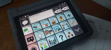 L'image montre une tablette avec une application de communication améliorée et alternative (CAA), affichant des symboles visuels et du texte pour différentes catégories comme la nourriture, l'école et les sentiments.