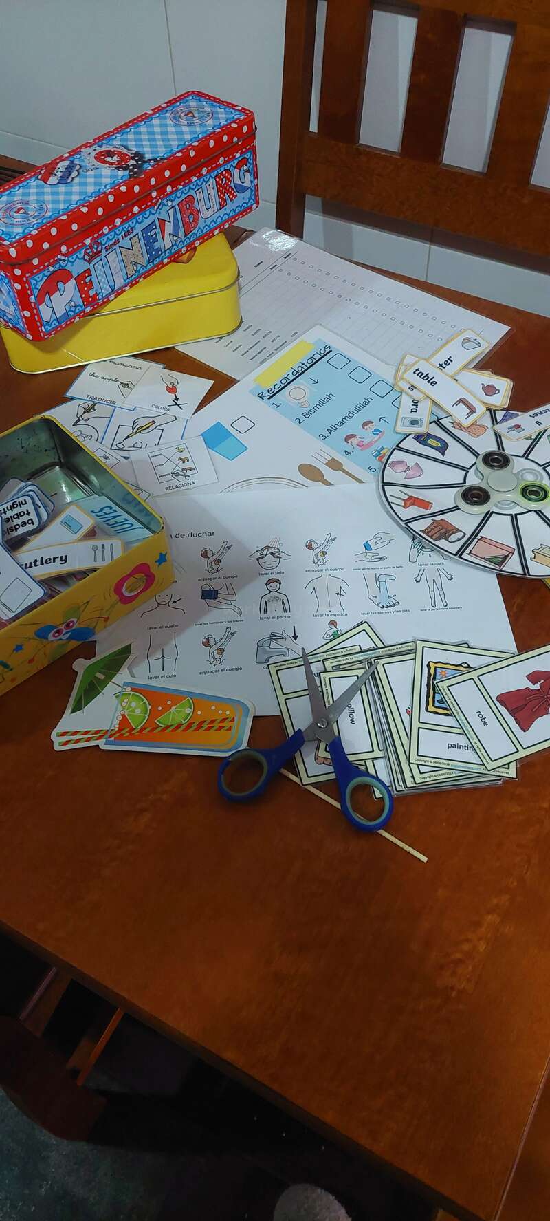 Cette image montre du matériel éducatif, notamment des cartes illustrées, des feuilles de travail, une toupie, des étiquettes de mots et des ciseaux, organisé pour une activité linguistique ou d'apprentissage amusante et interactive.