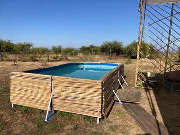 Ein kleiner, rechteckiger, oberirdischer Pool wird von Bambusplatten umschlossen und liegt draußen auf einer trockenen, grasbewachsenen Fläche mit vereinzelten Büschen und blauem Himmel.