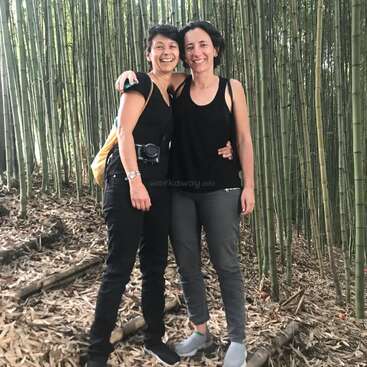 Two women stand together in a bamboo forest, embracing, with one wearing a camera around her waist and the other with her arm around her partner\'s shoulder.