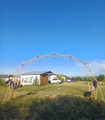 Zwei Menschen hängen kopfüber an einer Bambuskonstruktion auf einer Wiese. Im Hintergrund sind kleine Gebäude, geparkte Autos und ein klarer blauer Himmel zu sehen.