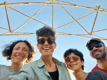 Vier lächelnde Menschen mit Sonnenbrillen posieren gemeinsam unter einer geometrischen Holzstruktur an einem sonnigen Tag mit blauem Himmel und fröhlichen Gesichtern.