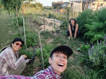 Drei lächelnde Menschen gärtnern zusammen in einem üppigen, grünen Garten. Sie sind von verschiedenen Pflanzen umgeben, genießen die freie Natur und wirken glücklich und entspannt.