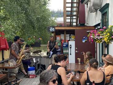 L'image représente un cadre extérieur animé, avec un groupe se produisant sur une terrasse, entouré de personnes assises à des tables, au milieu d'une verdure luxuriante et de fleurs éclatantes.