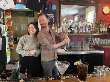L'image représente un homme et une femme debout derrière un bar, l'homme tenant un shaker et la femme portant un sweat-shirt gris. Le bar est bien approvisionné en boissons et en verres.