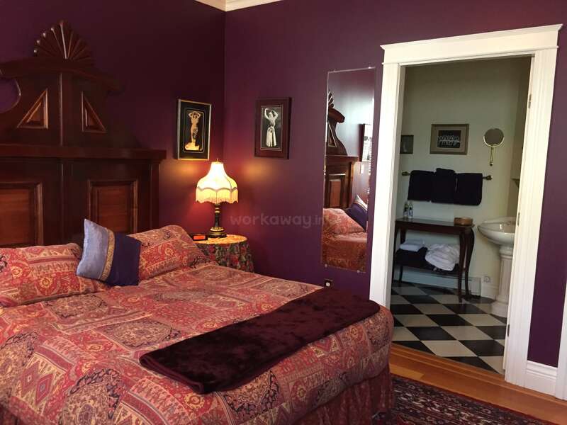 L'image représente une chambre à coucher avec un lit, une table de nuit, une lampe et un miroir, ainsi qu'une salle de bains avec un lavabo, des toilettes et un sol à carreaux, le tout sur un fond mauve.