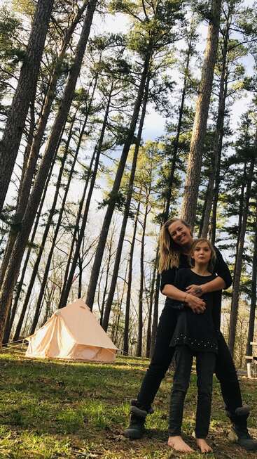 Une femme et une jeune fille sourient ensemble dans un camping en forêt, avec de grands pins et une tente beige en arrière-plan, profitant de la nature.