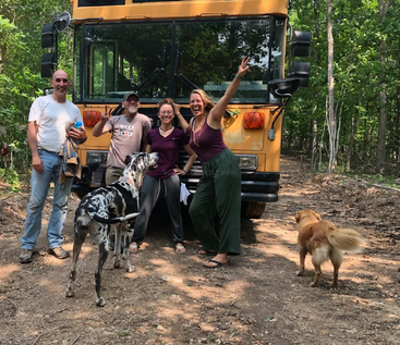 Quatre personnes souriantes et deux chiens posent devant un bus scolaire jaune sur un chemin de terre, entouré d'arbres verts, profitant d'une journée ensoleillée en plein air.