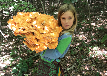 Une jeune fille se tient dans une forêt ensoleillée, tenant fièrement un gigantesque champignon orange vif, entourée d'arbres et de sous-bois feuillus, et portant une tenue colorée.