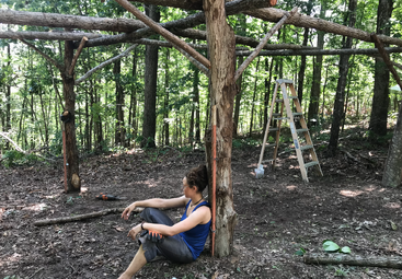 Une femme en tenue de travail de plein air s'appuie sur un poteau en bois au milieu d'une structure en rondins partiellement construite dans une forêt, avec une échelle et des outils à proximité.