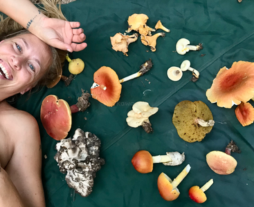 Une femme souriante est allongée sur une bâche verte entourée d'un assortiment de champignons sauvages de différentes formes, tailles et couleurs, créant ainsi une scène joyeuse inspirée de la nature.