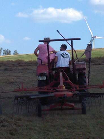 La imagen muestra a dos personas en un tractor rojo en un campo, con un aerogenerador visible al fondo bajo un cielo azul con nubes blancas.