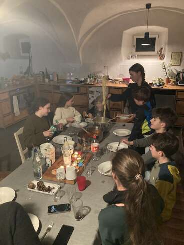 Eine Gruppe von sieben Leuten, hauptsächlich Kinder, sitzt um einen Küchentisch herum, während eine Frau Nudeln serviert. Die Küche wirkt gemütlich, warm und wohnlich.