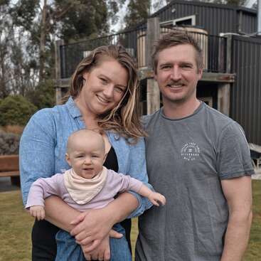 A happy family stands outdoors: a woman holding a baby, smiling warmly beside a man. Behind them, there are trees, grass, and a modern house.