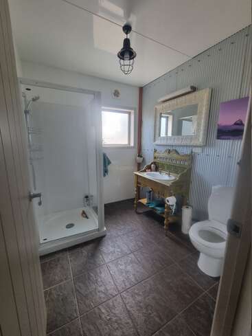 This bathroom features a glass shower, toilet, ornate mirror, rustic vanity, corrugated metal wall, dark tile floor, pendant light, window, and a purple landscape picture.