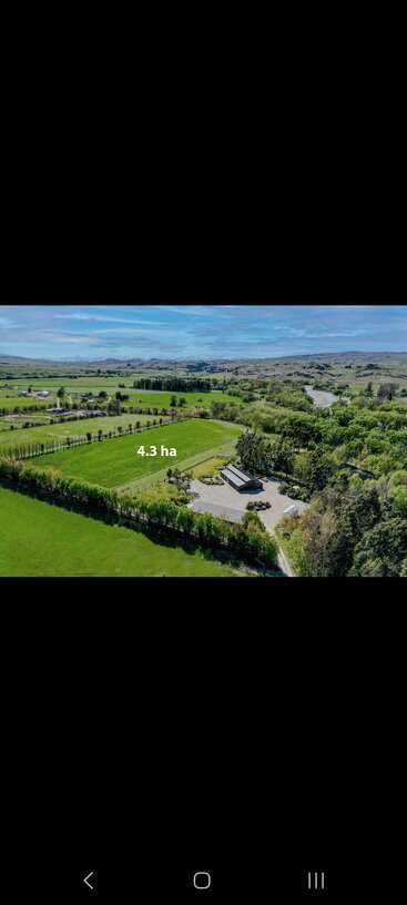 Luftaufnahme eines üppig grünen 4,3 Hektar großen Grundstücks mit eingezäunten Pferdekoppeln, einer Scheune, baumbestandenen Grenzen, Hügeln, einem Fluss und blauem Himmel im Hintergrund.