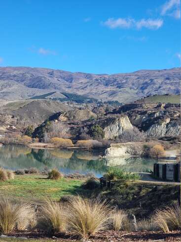 Eine heitere Landschaft mit einem ruhigen, spiegelnden See, schroffen Hügeln, spärlichen goldenen Bäumen, trockenem Gras und fernen Bergen unter einem strahlend blauen Himmel mit vereinzelten Wolken.