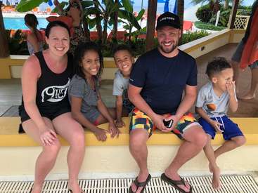 The image depicts a family of five sitting on a yellow bench, with a pool and tropical plants visible in the background.