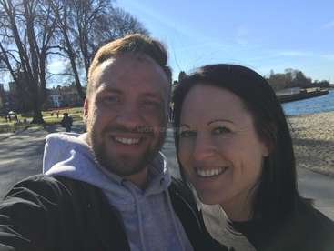 The image shows a selfie of a man and woman standing together in front of a park, lake, and trees on a sunny day.