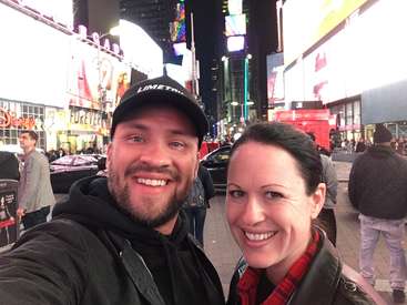 The image shows a selfie of a man and woman in Times Square, New York City, with a busy street and billboards in the background at night.