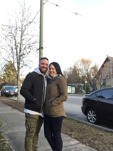 The image depicts a man and woman standing on a sidewalk, with a street, cars, and buildings visible in the background, set against an overcast sky.