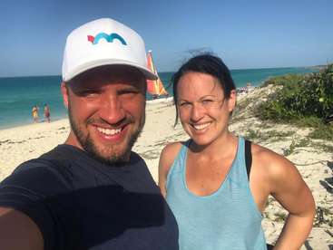 La imagen muestra a un hombre y una mujer tomándose un selfie en una playa, con el océano y un velero visibles al fondo.