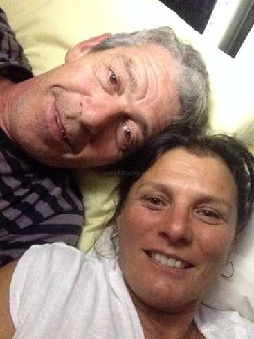 La imagen muestra un selfie de un hombre y una mujer tumbados en una cama, con la mujer vestida con una camisa blanca y el hombre con una camisa de rayas negras y grises.