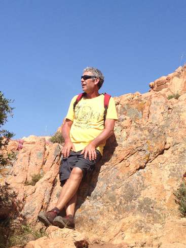 La imagen muestra a un hombre sentado en una roca, vestido con una camiseta amarilla, pantalones cortos negros y zapatillas de deporte, con una mochila roja al hombro y un cielo azul despejado.