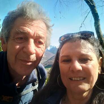 Un homme et une femme âgés sourient à l'extérieur en prenant un selfie. La lumière du soleil illumine leurs visages. Derrière eux, les montagnes, les arbres et le ciel bleu créent un arrière-plan pittoresque et joyeux.