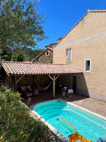 Une charmante arrière-cour avec une piscine étincelante, des jouets gonflables, un coin salon ombragé avec un hamac, un patio en pierre, une végétation luxuriante, une maison rustique et un ciel d'un bleu éclatant.