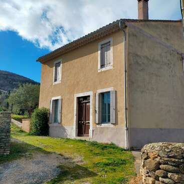 Une charmante maison beige à deux étages, avec des fenêtres à cadre blanc et une porte en bois foncé, située le long d'un chemin herbeux, sous un ciel bleu parsemé de nuages.