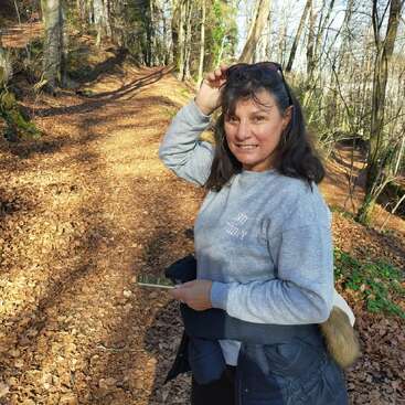 Une femme souriante se tient sur un sentier forestier ensoleillé couvert de feuilles mortes, tenant des lunettes de soleil et un téléphone, portant un pull gris et une veste autour de la taille.