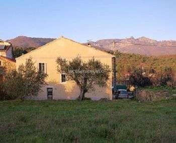 La imagen representa una casa amarilla con tejado a dos aguas, situada en un entorno rural con un árbol en primer plano y una cadena montañosa al fondo.