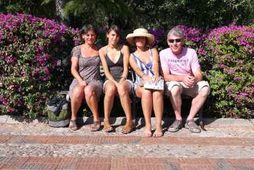 La imagen muestra a tres personas sentadas en un banco, rodeadas de un seto de flores moradas, con árboles y un camino de piedra como telón de fondo.