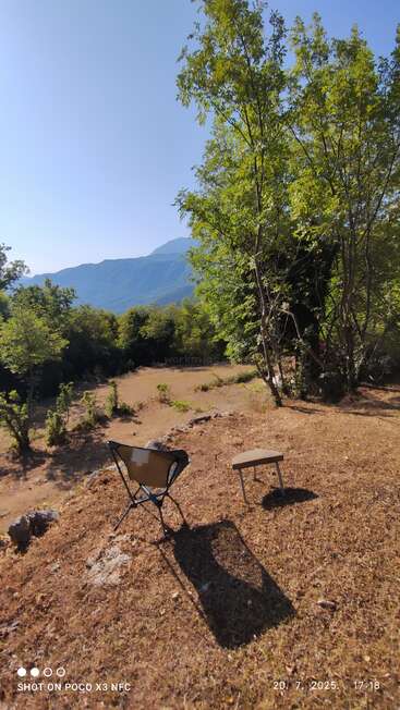 A solitary chair and small table sit on dry grass, surrounded by green trees and mountains under a bright blue sky, radiating peace and solitude.