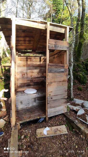 This image shows a rustic wooden outdoor toilet structure in the woods. The door is open, revealing a makeshift seat. Leaves and debris surround it.