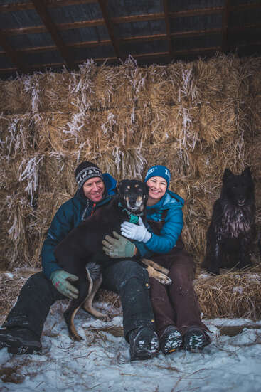 Um casal feliz está sentado junto em fardos de feno, bem agasalhado com roupas de inverno, sorrindo ao lado do seu cachorro preto, enquanto outro cachorro preto e fofinho está sentado ali perto, na neve.