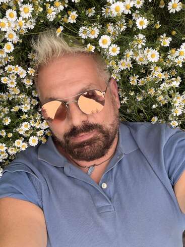 Ein Mann mit blonden Haaren und Bart liegt auf Gänseblümchen, trägt eine rosa getönte Sonnenbrille und ein blaues Poloshirt und genießt einen friedlichen, sonnigen Moment im Freien.