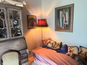 Ein gemütliches Zimmer mit einer Couch mit kunstvollen Kissen, einem roten Lampenschirm, abstrakten Wandbildern und Motiven mit Tieren, einem Vintage-Schrank und einem bequemen Sessel. Warme Beleuchtung.