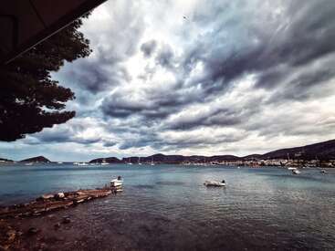 Unas nubes oscuras y amenazantes se ciernen sobre una bahía tranquila con barcos fondeados. Las aguas azules y tranquilas contrastan con el cielo sombrío. Las montañas lejanas y un pueblo costero completan la escena.