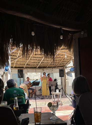 A live band performs on a small stage in a thatched-roof venue. Audience members watch while enjoying drinks, creating a relaxed, tropical atmosphere.